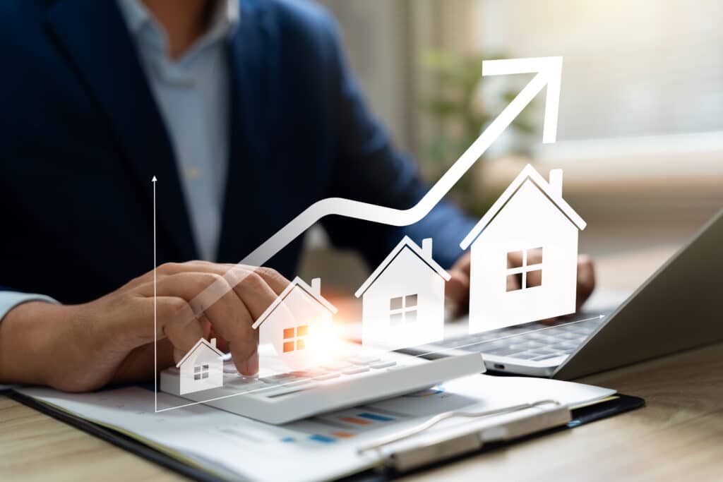 Main sur une calculatrice avec des maisons de tailles croissantes et une flèche ascendante symbolisant l'augmentation de la valeur immobilière et l'investissement.