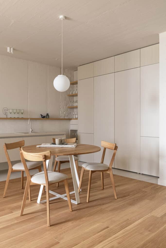 Cuisine et salle à manger minimalistes avec table ronde en bois, chaises claires, suspension moderne. Placards intégrés et sol parquet.