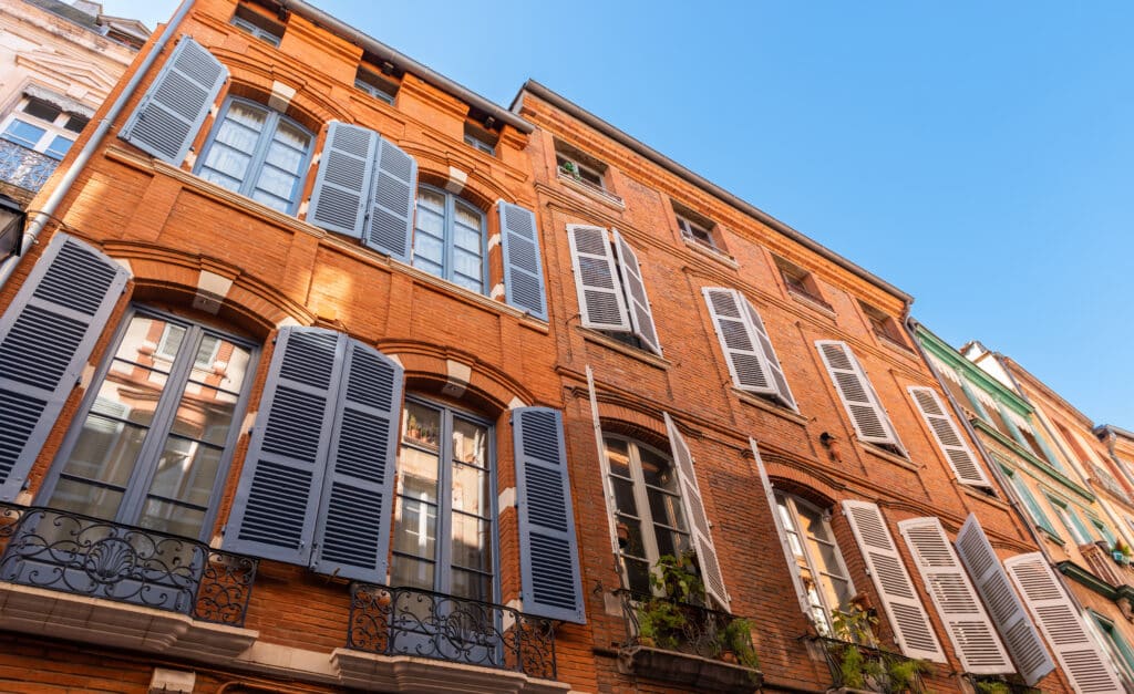 Façade en brique rouge d'un immeuble toulousain avec fenêtres à volets bleus et blancs ouverts et balcons sous ciel clair.
