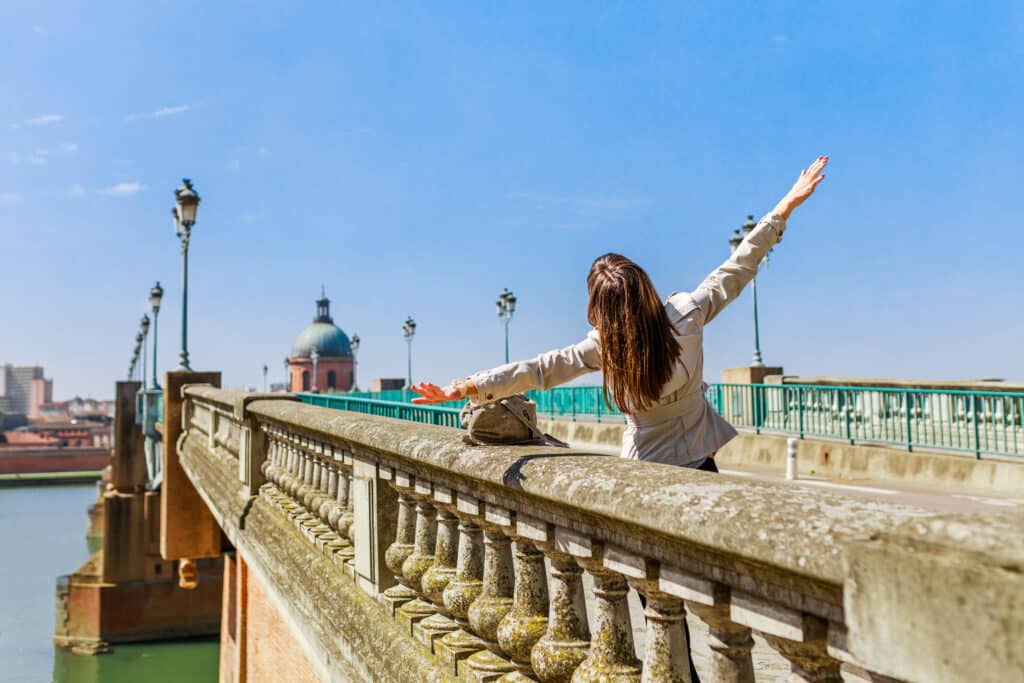 Femme les bras ouverts sur un pont en pierre. Vue sur le dôme de la Grave de Toulouse et la Garonne sous ciel bleu.